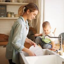 Woman doing dishes while baby sits on counter watching.